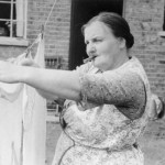 Eine Hausfrau mit einer Wäscheklammer zwischen den Lippen hängt ein weißes Stück Wäsche zum Trocknen auf.